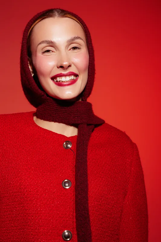 Smiling woman in red woolen coat and matching scarf poses against red background. Bright makeup and joyful expression create festive and warm mood for winter fashion concept.