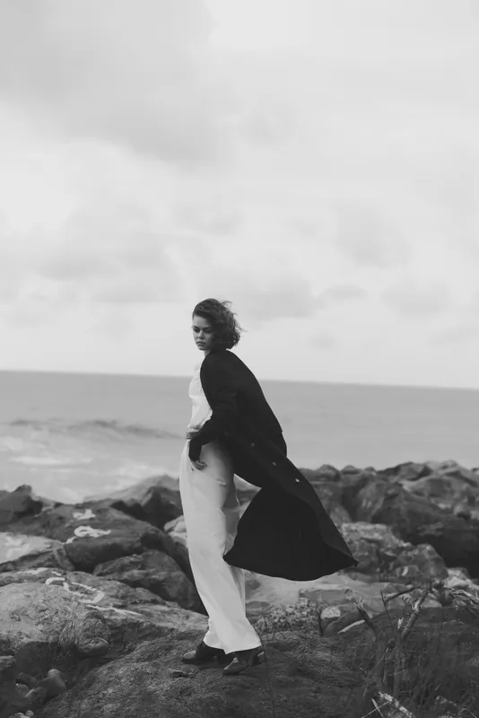 Black and white photo of a woman in a flowing coat on a rocky coastline, wind in her hair, ocean in the background, moody fashion scene by the sea