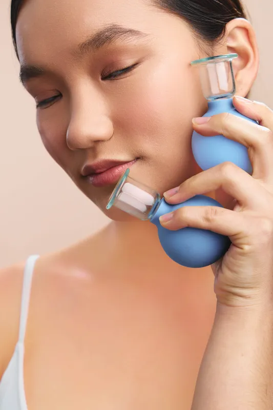 Asian woman uses a facial vacuum device in a calm home setting, highlighting self care and wellness. She smiles softly, conveying relaxation and focus while embracing modern beauty technology for