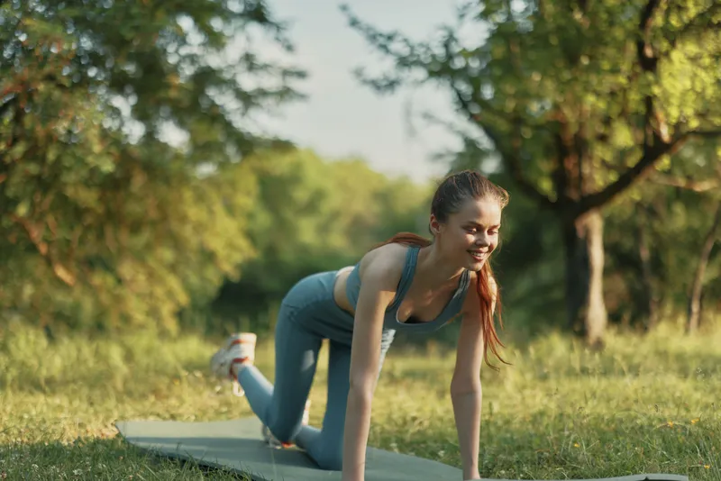 Young athletic woman exercising outdoors in a park, wearing a blue sports outfit, showcasing joy and fitness on a sunny day, surrounded by green trees