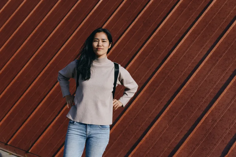 Woman standing with hands on hips against an angled wooden wall, casual sweater and jeans, backpack strapped, urban outdoor vibe, natural light, confident pose for everyday style.