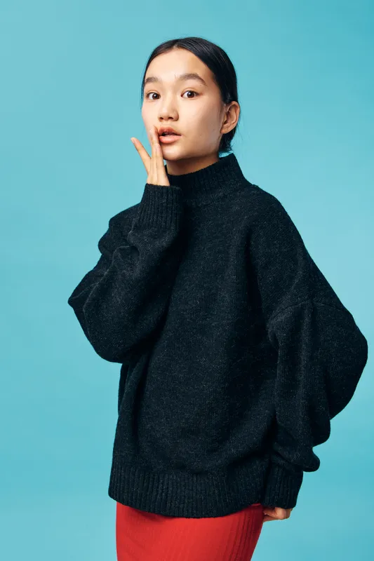 Woman wearing black sweater and red skirt standing against blue background with thoughtful expression and hand near face, casual style and studio portrait with soft lighting.
