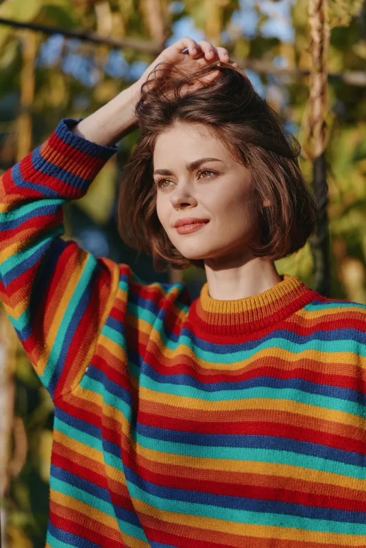 Woman portrait in colorful striped rainbow sweater outdoors, sunlight on face, short hair and touching hair with hand, natural expression and warm autumn background for lifestyle fashion.