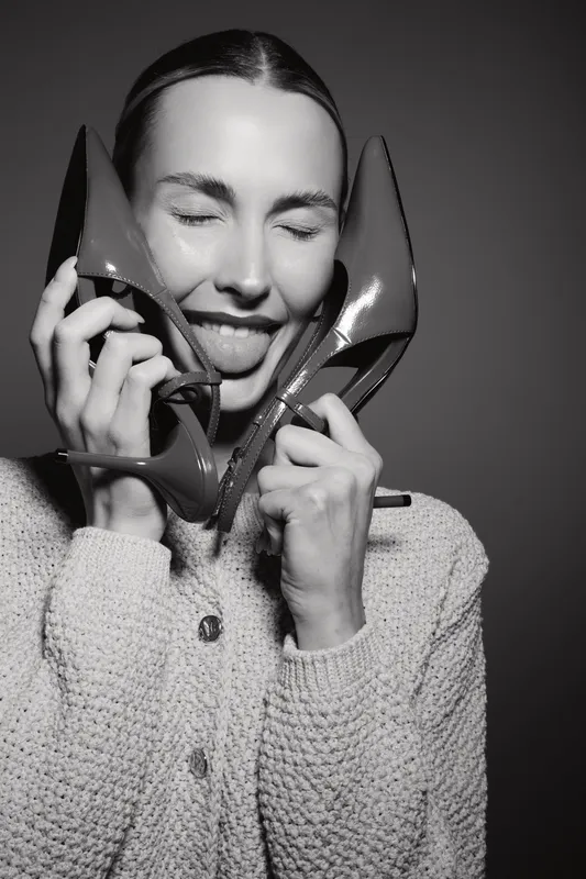 Young Caucasian woman with light skin playfully holds high heel shoes near her face with closed eyes and tongue out. Concept of fun, fashion, and lifestyle in studio black and white.