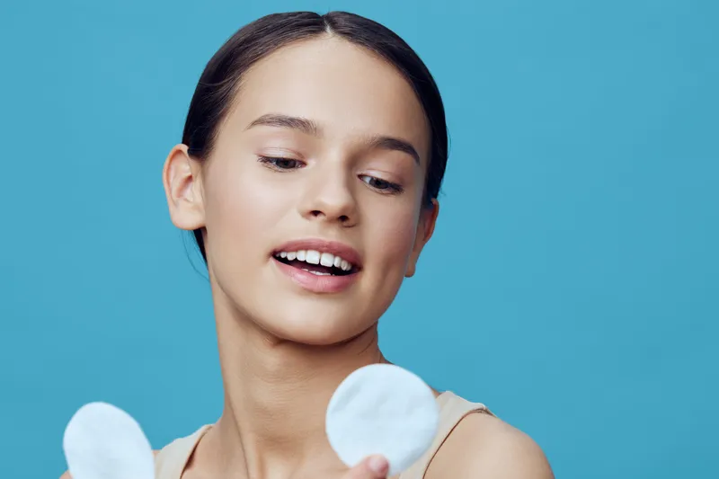 Young woman with clear skin holding cotton pads, smiling against a blue background, representing skincare routine and self care concept