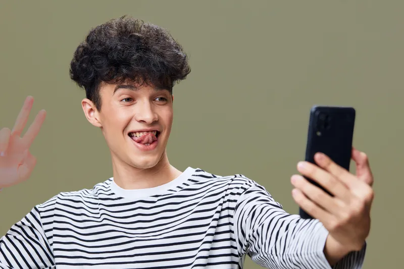 young man taking selfie with smartphone, waving hand and playful expression, casual striped shirt, light green background, indoor portrait