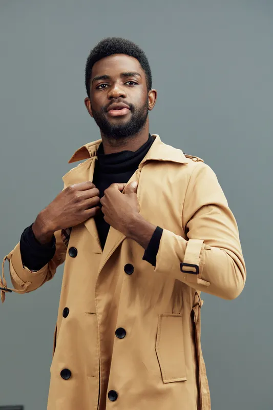 Stylish African man in a beige trench coat, exuding confidence against a muted gray background, ideal for fashion and autumn themed projects