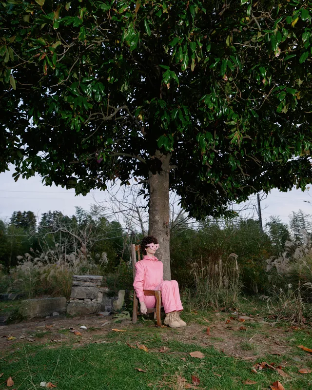 Woman in a pink dress sits on a chair beneath a leafy tree in an outdoor garden setting, exuding calm and serene mood amid rustic surroundings and natural light.