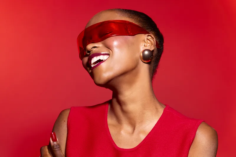 Smiling black woman with dark skin wearing red sleeveless top and red futuristic sunglasses against red background. Concept of fashion, style, confidence, and modern beauty.