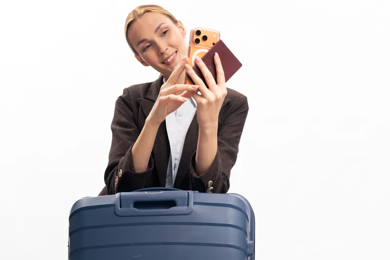Young woman with suitcase holds passport and smartphone taking selfie on white background. Concept of travel, vacation, tourism, and modern lifestyle with happy female traveler.