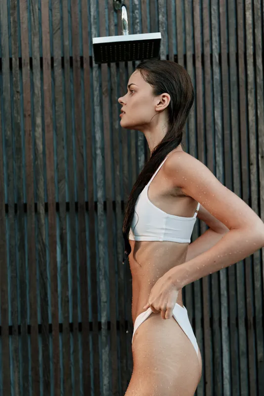 A fit woman stands by an outdoor shower in a white swimsuit, water droplets glistening on her skin. A lifestyle moment highlighting fitness, wellness, and outdoor routine.