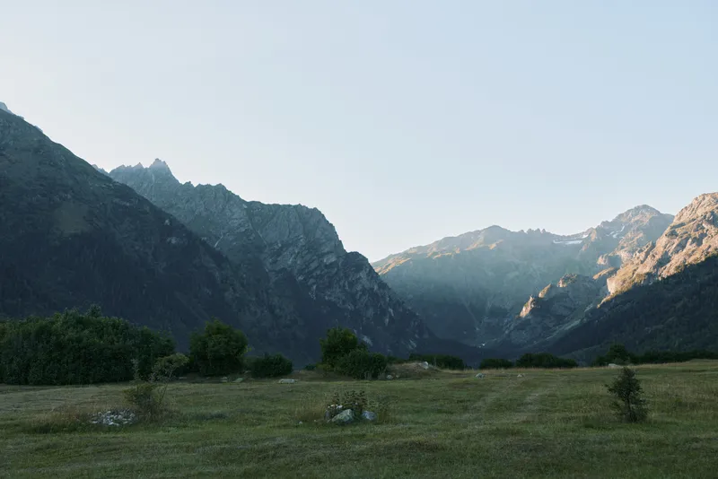 A serene mountain valley opens to a wide grassy plain beneath a clear sky, with rugged peaks in the distance, inviting calm, exploration, and peaceful reflection.