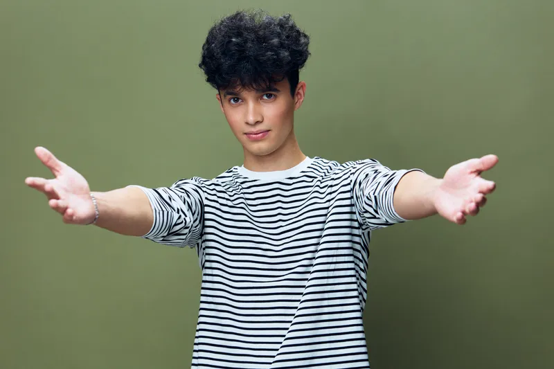 Young man with curly hair wearing a striped shirt stretches arms forward against green background, expressing openness and warmth in casual style.