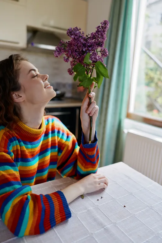 Woman sits at a kitchen table in a colorful striped sweater, smelling a lilac bouquet, enjoying a cozy home moment, bright window light, relaxed and content.