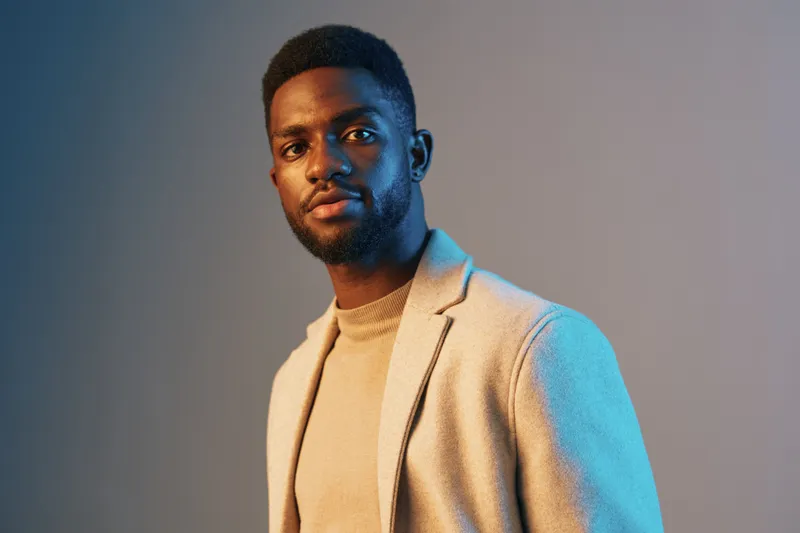 Stylish young Black man in a beige blazer and sweater against a soft blue gradient background, exuding confidence and charisma for fashion and lifestyle imagery