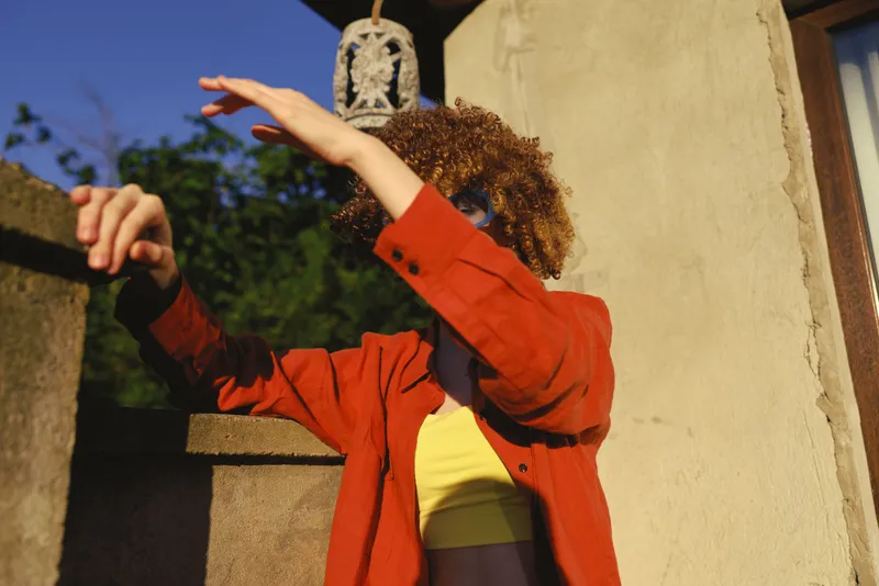Joyful woman outdoors in a bright red jacket and yellow top, mid pose with outstretched arm, sunlit moment, natural colors, relaxed expression, warm breeze and bright backdrop setting