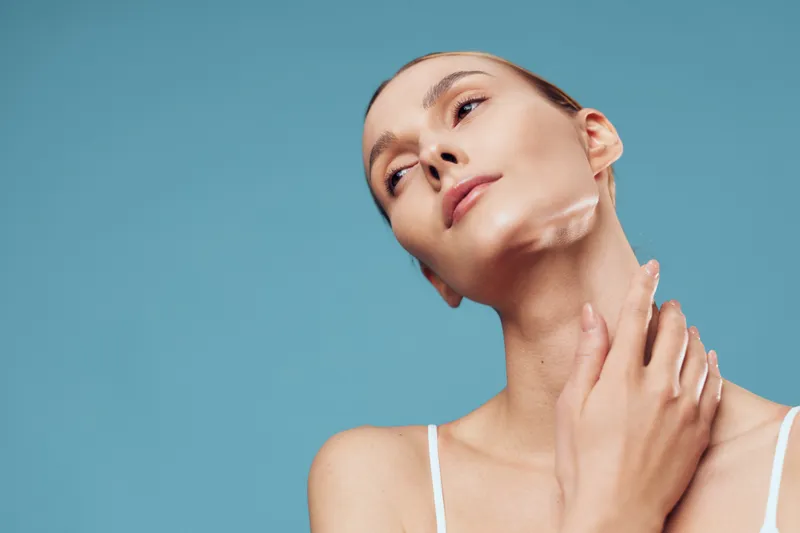 A young woman tilts her head back slightly and touches her neck in a studio with a blue background, conveying calm self care and quiet confidence.