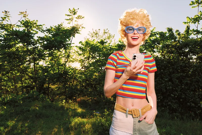 Woman in striped crop top and shorts poses outdoors in a sunny park, taking a selfie for a retro fashion portrait, casual lifestyle vibe, cheerful and confident.