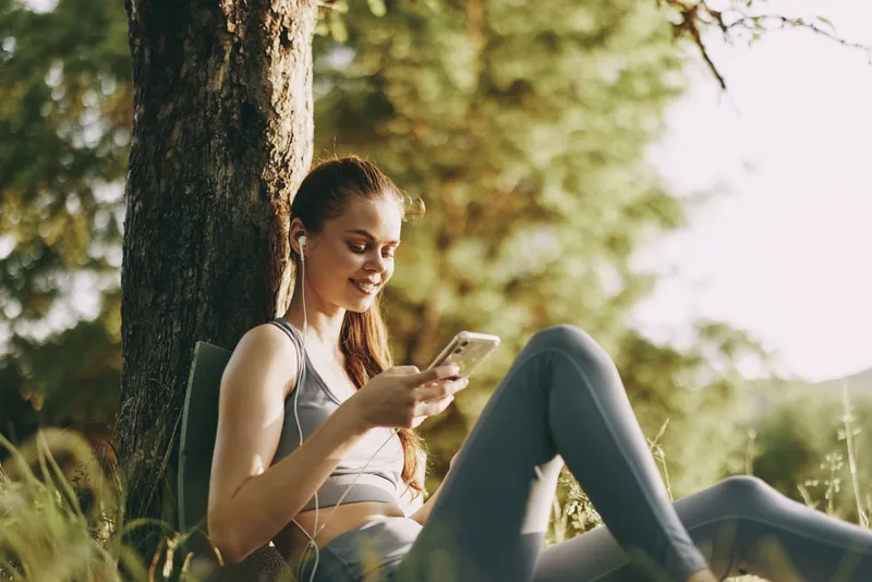 Young woman enjoying music on her smartphone outdoors, wearing activewear and sitting by a tree, conveying relaxation and joy in nature
