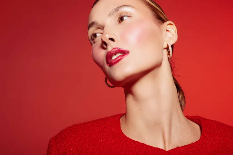 Portrait of a stylish woman with red lipstick and earrings wearing a red sweater against a red background. Concept of fashion, beauty, elegance, and confident attitude.