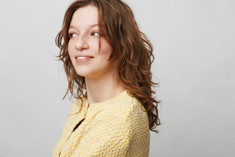 Young woman with curly brown hair wearing a yellow sweater looks sideways with a slight smile. Studio portrait on a light grey background. Concept of natural beauty and casual style.