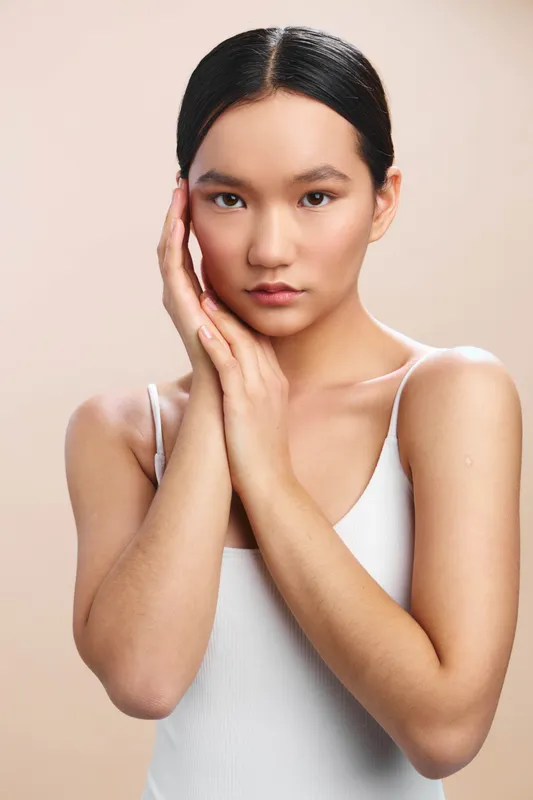 Asian woman portrait in a studio, calm expression, hands framing face, clean neutral backdrop, soft lighting highlights smooth skin, poised and thoughtful mood, professional studio headshot vibe.