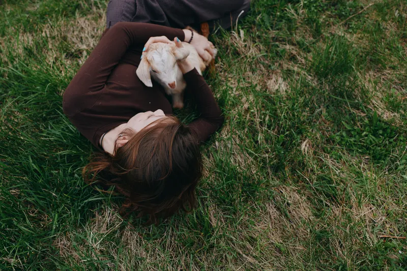 Calm lifestyle scene shows a woman resting on lush grass, embracing a playful goat kid, capturing a gentle bond with animals, peaceful outdoor moment, personal connection and rural serenity