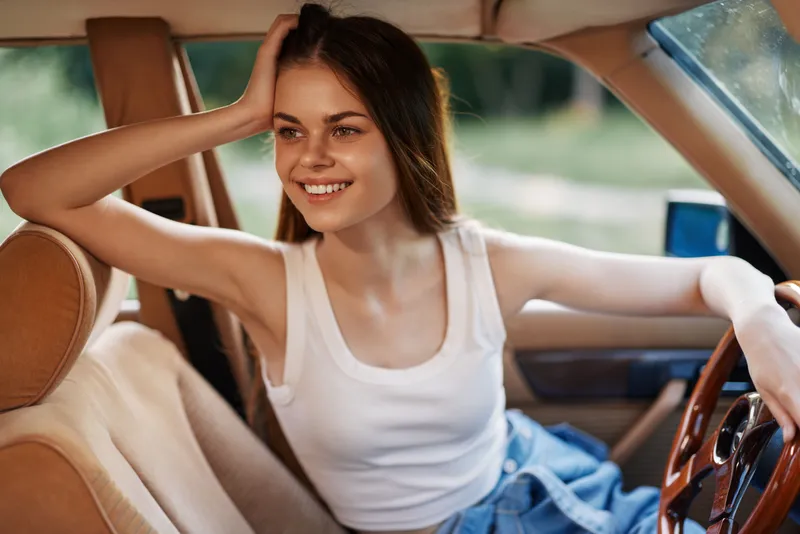 Young woman smiling in a car, wearing a white tank top and denim shorts, enjoying a sunny day, showcasing a summer vibe and happiness on her face.