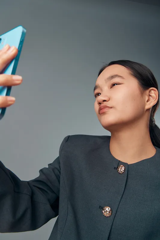 Young woman taking selfie with smartphone in business attire on grey background. Casual pose with confident look, modern technology usage and communication in portrait style.