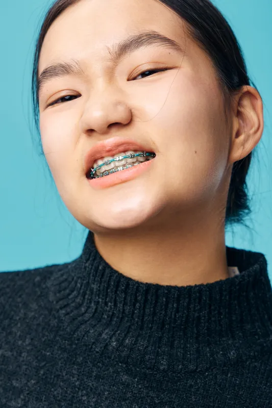 Closeup portrait of young woman wearing braces with metal brackets smiling showing teeth against blue background. Person with dental orthodontic treatment and confident facial expression in casual