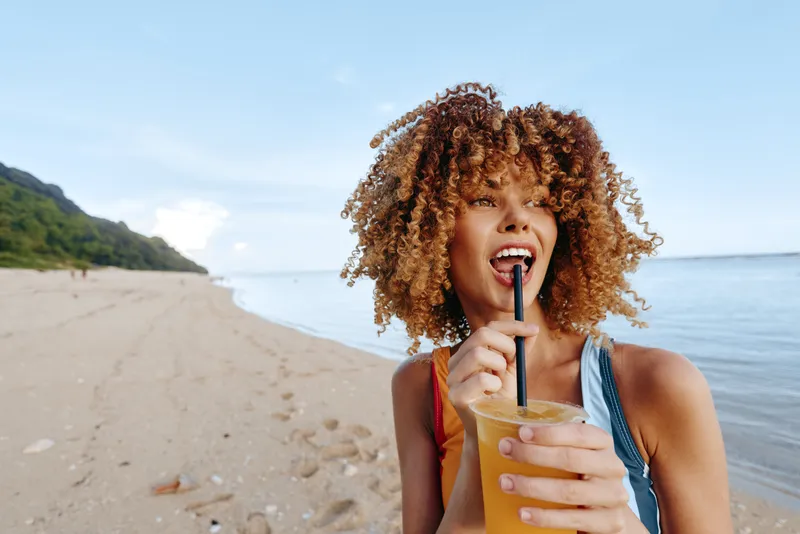 Young woman with curly hair enjoys a cold drink through a straw while standing on a sandy beach near calm water under a clear blue sky.
