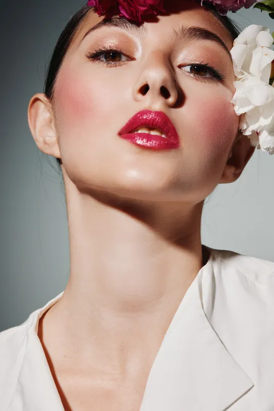 Porcelain skin woman with bright glossy lipstick and soft blush poses close-up. Floral accessories emphasize beauty and fashion with creative makeup styling on visible face.