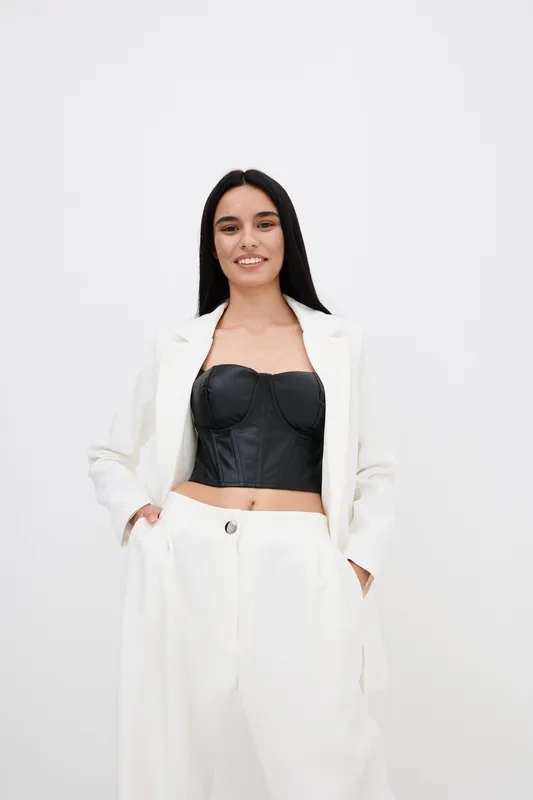 woman in white suit wearing black corset top smiling confidently with hands in pockets against plain background stylish modern look