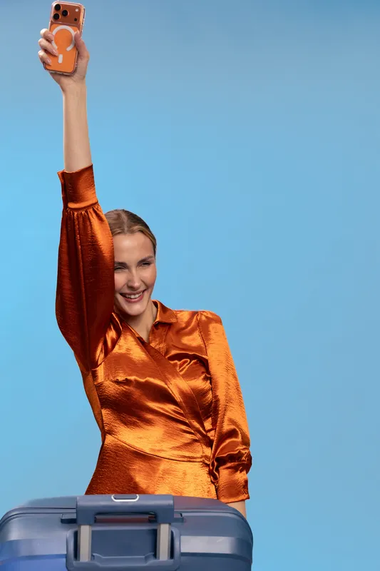 Smiling woman in shiny orange blouse holds smartphone high and poses with suitcase against blue background. Concept of travel, vacation, happiness, and modern lifestyle.