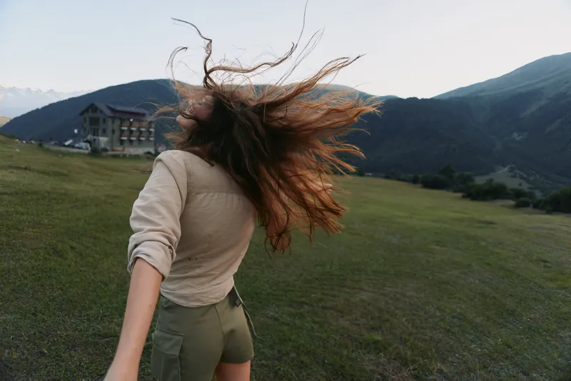 Young woman with long hair stands on green hill holding hand of partner looks back at mountain landscape during outdoor adventure in nature lifestyle concept