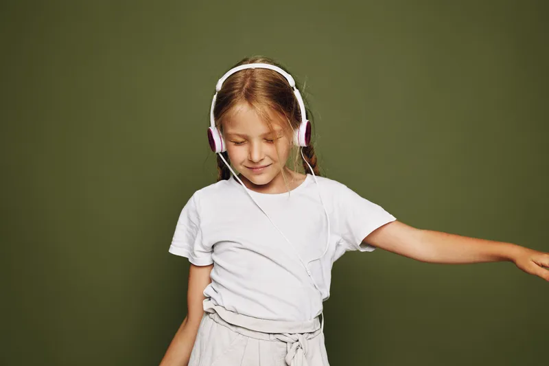 Happy young girl with headphones enjoys music with closed eyes, wearing a white t-shirt against a green background. Concept for childhood, relaxation, and music enjoyment.