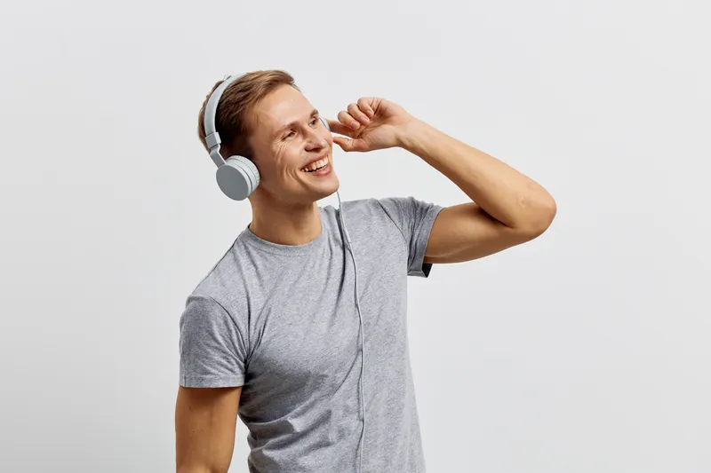 Man wearing headphones enjoying music, smiling, looking happy, casual grey t-shirt, minimalistic white background, studio portrait, lifestyle concept, cheerful and relaxed mood.