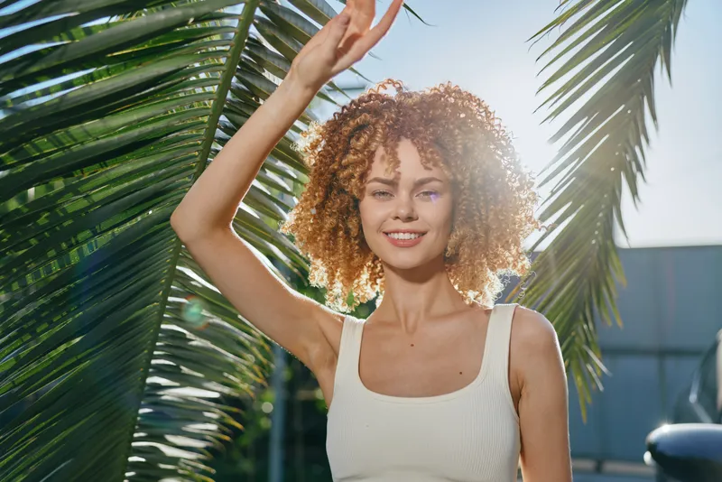 smiling woman with curly hair enjoys sunny weather outdoors near tropical palm leaves dressed in casual white top, natural light creating warm summer atmosphere