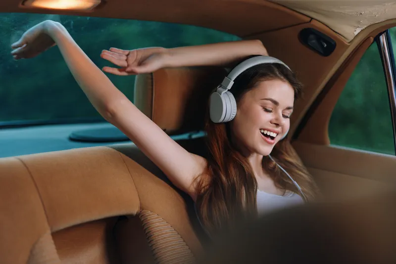 Young woman enjoying music with headphones, relaxing in a car, smiling with carefree emotions, showcasing a cozy and stylish environment.
