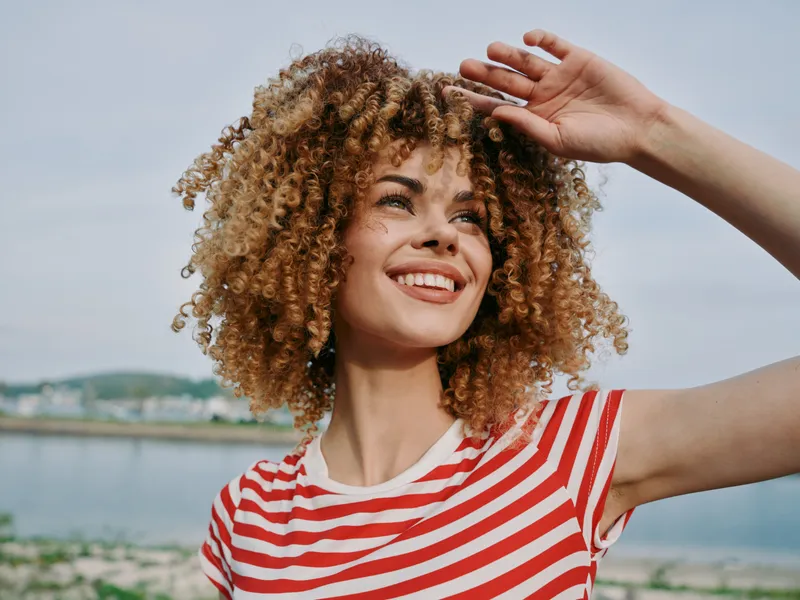 Smiling woman with curly hair stands outdoors by a calm water body, wearing a red and white striped shirt, sunlit face and vibrant summer vibe.