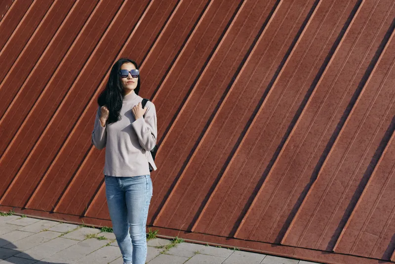 Stylish woman poses beside a diagonal wooden wall wearing sunglasses scarf and jeans showcasing casual street fashion and urban attitude in bright daylight