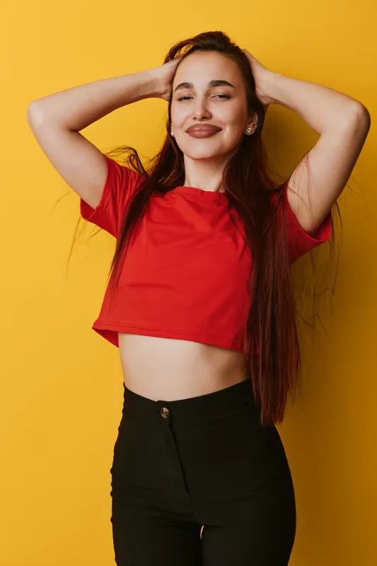Young woman with long hair posing with hands behind head smiling and wearing red crop top and black pants against yellow background in studio portrait.