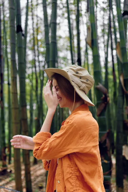 woman bamboo hat orange forest portrait, traveler in a wide brim hat and bright orange jacket walking through a bamboo grove, candid side profile smiling in soft natural light