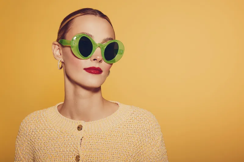Stylish woman with green round sunglasses and red lipstick poses against yellow background wearing beige knitted sweater. Concept of fashion, summer style, and confident attitude.