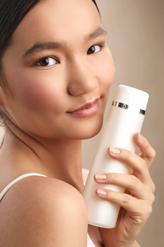asian woman holds a white skincare bottle close to her face, smiling softly in warm studio light, conveying gentle beauty, self care, and calm confidence in a serene portrait.
