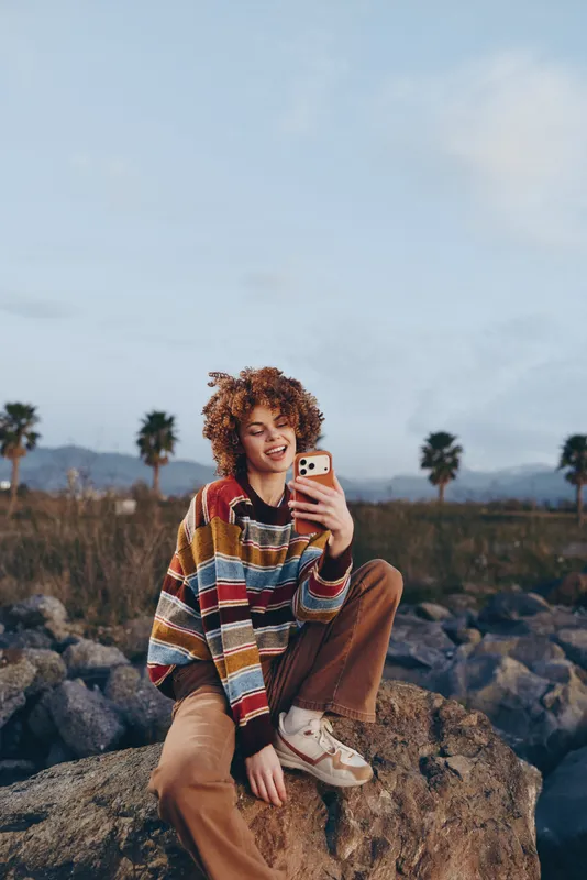 Woman lifestyle outdoors selfie smartphone casual scarf rocks at sunset, sitting on stone, warm light, relaxed smile, palm trees, distant mountains, candid moment, breezy sky, calm