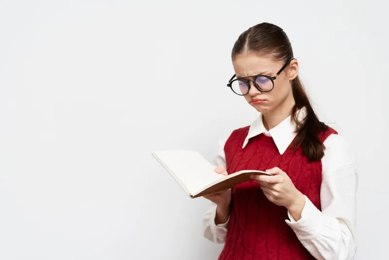 Молодая девушка в очках и красном свитере читает книгу с озадаченным выражением лица