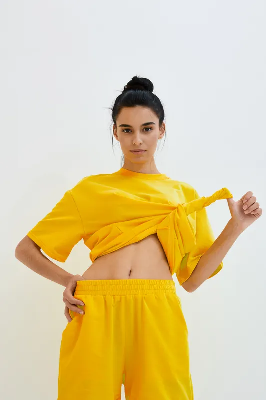 young woman in yellow casual outfit knots loose top, showing midriff confidently against plain light background, minimalistic style and relaxed pose