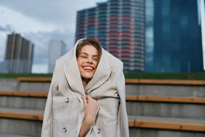 Woman wrapped in coat smiling happily outdoors urban background winter fashion style autumn season woman joy coat modern cityscape