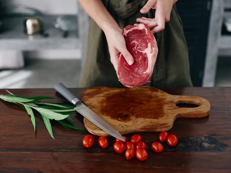 Женщина с ножом в руке нарезает свежий стейк из мяса для жарки на кухне